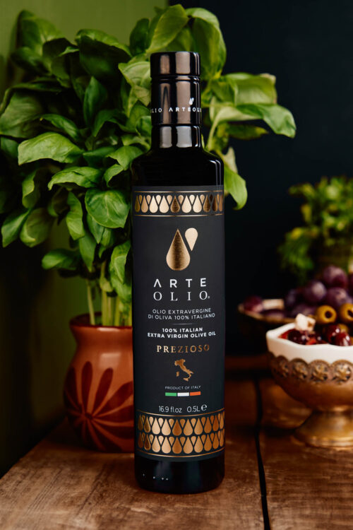 Arte Olio Extra Virgin Olive Oil Prezioso sitting on antique wood table with plants and olives in background.