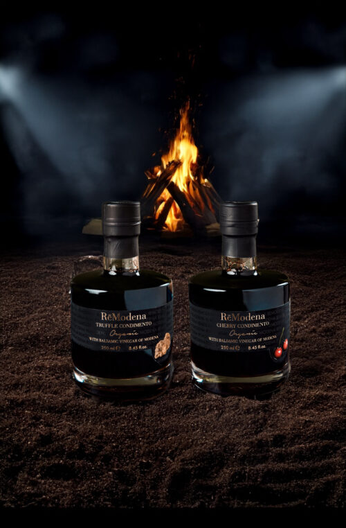 Two bottles of ReModena Balsamic sitting in sand in front of a campfire at night.