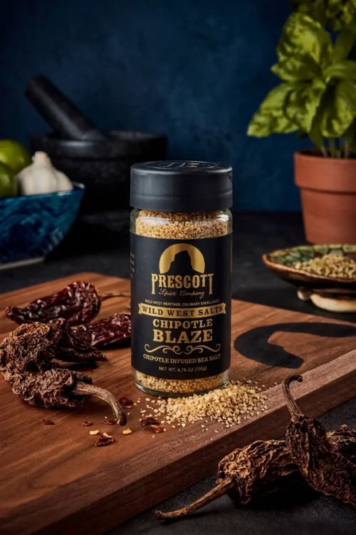 Prescott Spice Company Wild West Salts Chipotle Blaze Chipotle Infused Sea Salt glass jar on a wooden cutting board, surrounded by dried chipotle peppers, spilled salt crystals, a black mortar and pestle, and a potted basil plant in the background.