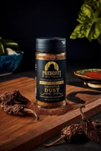 Prescott Spice Company Chipotle Dust Southwest Seasoning glass jar on a wooden cutting board, sitting on a bed of loose spice powder and surrounded by dried chipotle peppers, with a blurred potted basil plant in the background.