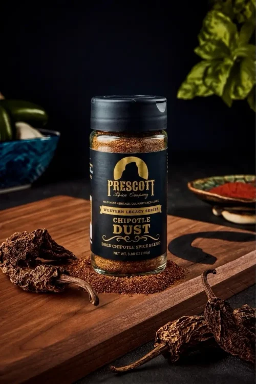 Prescott Spice Company Chipotle Dust Southwest Seasoning glass jar on a wooden cutting board, sitting on a bed of loose spice powder and surrounded by dried chipotle peppers, with a blurred potted basil plant in the background.