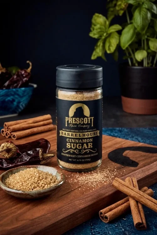 Prescott Spice Company Barrelhouse Southwest Cinnamon Sugar Southwest Cinnamon Sugar glass jar on a wooden cutting board, surrounded by cinnamon sticks, dried red chili peppers, a small ceramic bowl of brown sugar crystals, and a blurred basil plant in the background.
