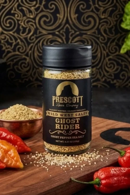 Prescott Spice Company Wild West Salts Ghost Rider Ghost Pepper Sea Salt glass jar on a wooden cutting board, surrounded by fresh red and orange ghost pepper chiles, a small copper bowl of spicy sea salt, and a black patterned backdrop.