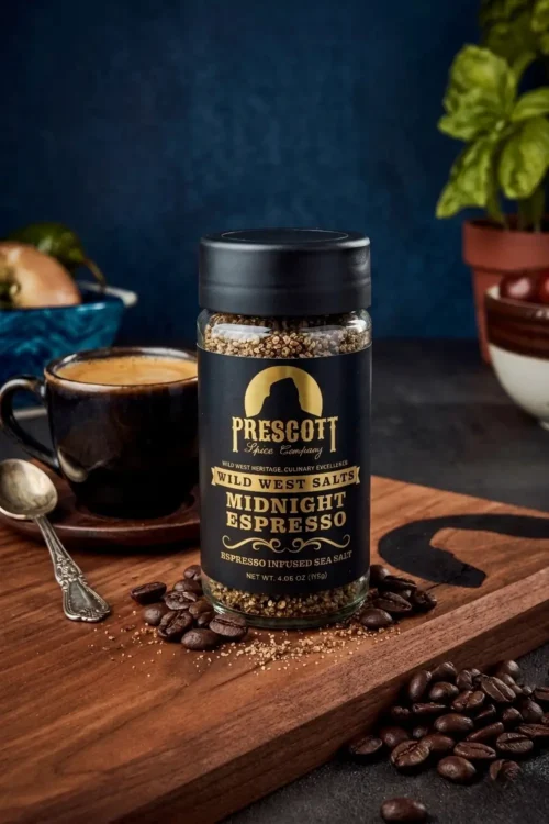 Prescott Spice Company Wild West Salts Midnight Espresso Espresso Infused Sea Salt glass jar on a wooden cutting board, with roasted coffee beans, a cup of fresh espresso with a spoon, and a blurred potted basil plant in the background.