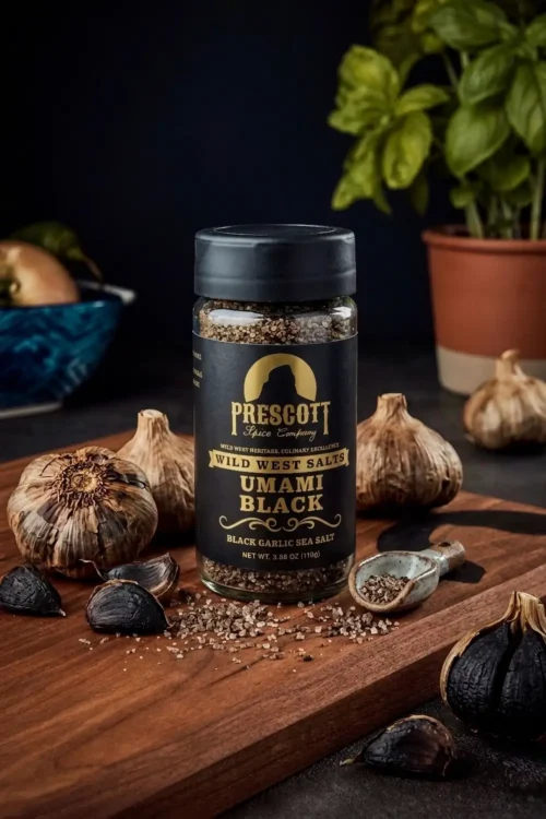 Prescott Spice Company Umami Black Garlic Sea Salt glass jar on a wooden cutting board, surrounded by whole bulbs and cloves of black garlic, a small ceramic scoop with spilled salt grains, and a potted basil plant in the blurred background.
